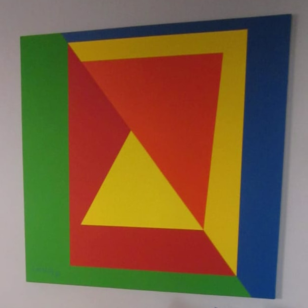 Obra geométrica de borde verde y azul con un recuadro amarillo, un plano rojo y un triángulo amarillo, colgada en una pared blanca.